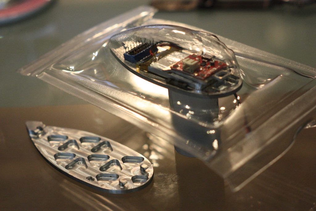 A circuit board mounted on an aluminum machining covered by a transparent piece of vacu-formed plastic, untrimmed.